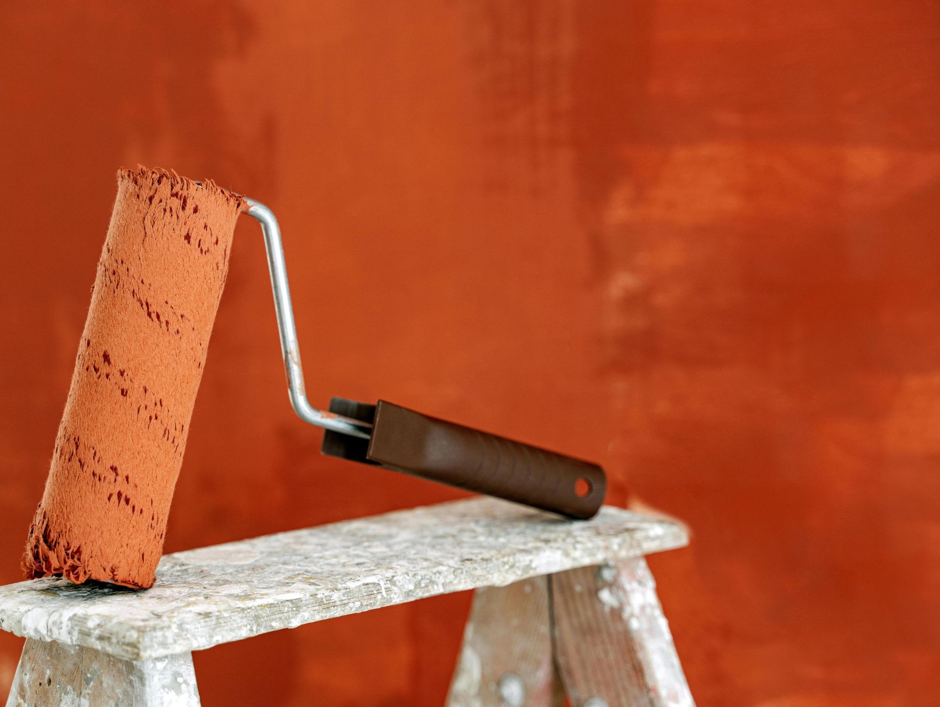 Detailed image of a paint roller resting on a ladder with a vibrant orange wall in the background.